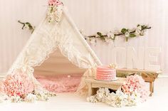 a teepee tent with pink and white flowers on the floor, next to it is a cake