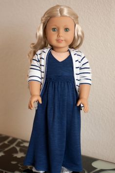 a doll with blonde hair wearing a blue dress and striped cardigan, standing in front of a white wall