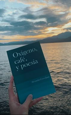 a person holding up a book in front of the water at sunset or sunrise with mountains and clouds behind them