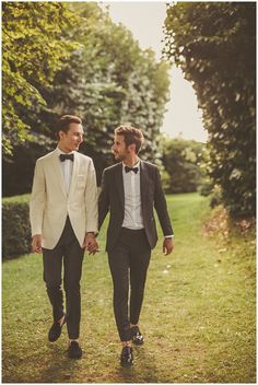 two men in tuxedos are walking down the grass holding each other's hands