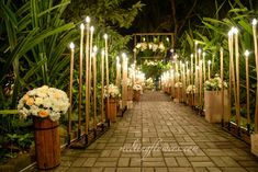 an outdoor walkway with candles and flowers on each side, surrounded by greenery at night