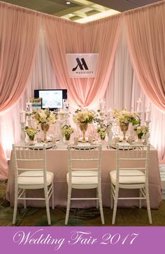 a table set up for a wedding with white chairs and pink drapes on the windows