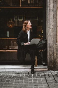 a woman sitting on a bench in front of a building using a laptop computer by an entrance