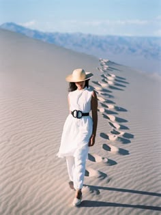 a woman in a white dress and hat walking on sand dunes