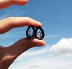 someone is holding two small black earrings in their hand, with the sky and clouds behind them