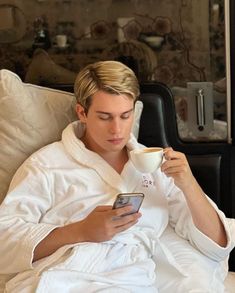 a man sitting on a couch holding a cup and cell phone in his hand while drinking from a mug