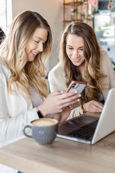 two women looking at their cell phones while sitting in front of a laptop computer on a table