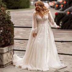 a woman in a long sleeved wedding dress is standing on the sidewalk near a fence