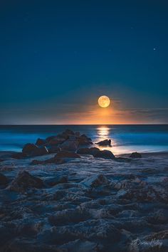 the full moon is setting over the ocean with rocks in the foreground and water below it