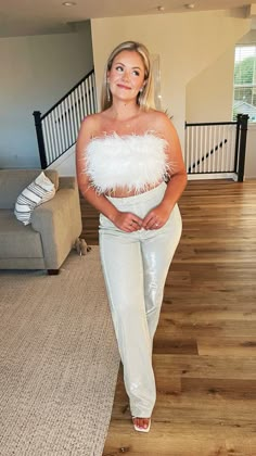 a woman standing in a living room wearing white pants and a top with feathers on it