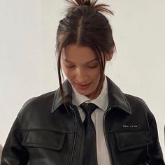 a woman wearing a black leather jacket and tie looking at her cell phone while standing in front of a white wall