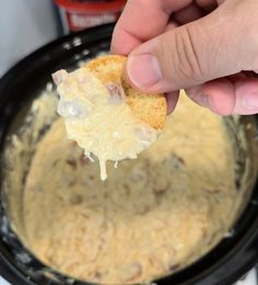 a hand holding a cracker over a crock pot filled with cheese and meat