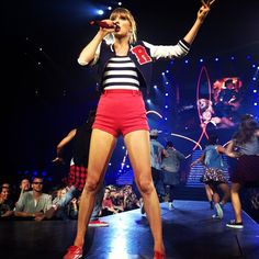 a woman in red shorts and striped shirt singing on stage with other people behind her