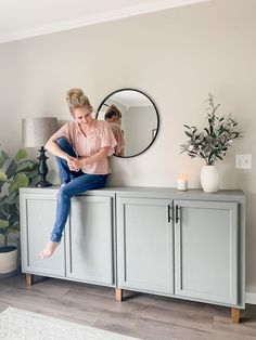 a woman sitting on top of a gray cabinet next to a mirror and potted plant