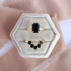 two rings with black stones are sitting on a white velvet ring box and one is in the shape of an octagon