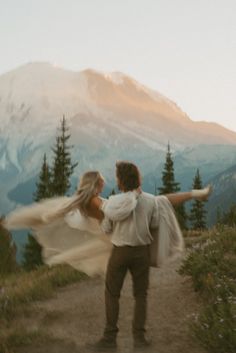 a man and woman holding each other on top of a hill with mountains in the background