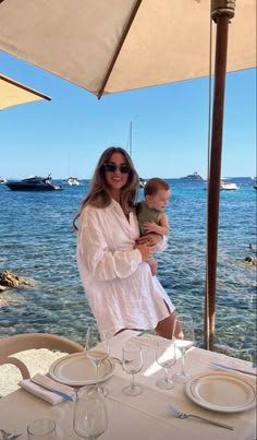 a woman holding a baby in her arms while sitting at a table with water and boats in the background