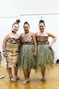 three women in grass skirts standing on a wooden floor with their arms around each other