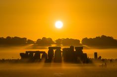 the sun is setting over stonehenge on a foggy day with people walking in the foreground