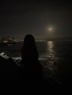 a woman sitting on the edge of a cliff looking out at the ocean with the moon in the distance