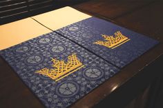 two blue and gold placemats sitting on top of a wooden table next to each other