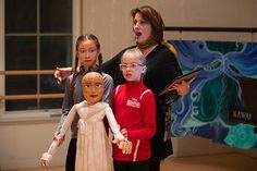 three girls and one boy are standing in front of a doll with an alien head on it