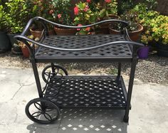 a black metal serving cart with wheels in front of some potted plants and flowers