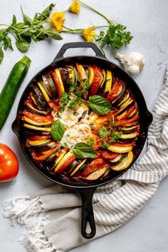 an iron skillet filled with vegetables on top of a white table next to a tomato and cucumber