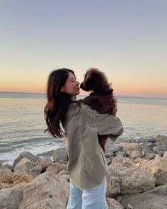 a woman standing on top of a rocky beach next to the ocean holding a dog