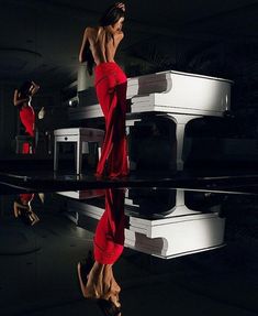 a woman in a red dress standing next to a white piano with its reflection on the ground