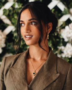 a woman in a brown jacket and gold necklace standing next to a wall with white flowers