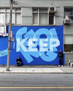 two people are sitting on the sidewalk in front of a blue wall that says keep