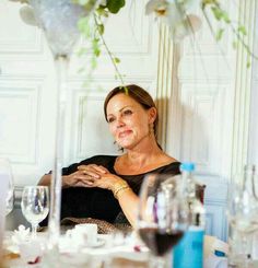 a woman sitting at a table with wine glasses and flowers in the corner behind her