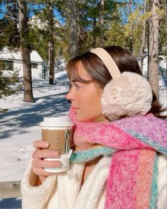 a woman wearing ear muffs holding a cup of coffee in the snow with headphones on