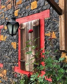 a red window with wooden shutters on the side of a stone building next to flowers