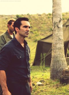 two men standing next to each other in front of a tree and tent on the ground