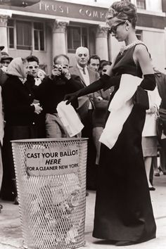 a woman standing next to a trash can filled with balls and papers while people look on