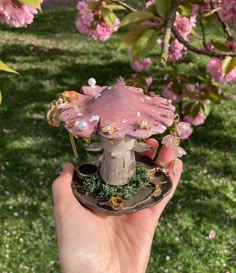 a hand holding a miniature fairy mushroom in front of some pink flowers and trees with lots of leaves