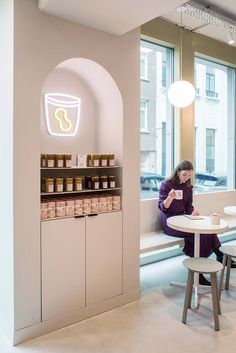 a woman sitting at a table in front of a window next to a shelf with jars on it