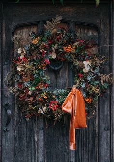 a wreath with an orange bow hanging on the front door to a wooden door decorated with greenery and foliage