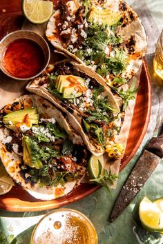 three tacos on a plate with salsa and limes next to it, along with two glasses of beer