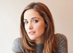 a woman with long brown hair wearing a sweater and looking off into the distance while sitting in front of a white wall