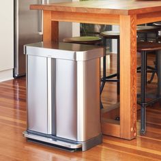 a stainless steel trash can sitting on top of a wooden floor next to a table