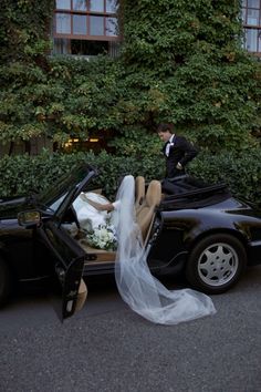 a bride and groom standing next to a black sports car with the doors open, in front of a green ivy covered building