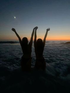 two women are standing in the water at sunset with their arms up to the sky