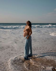 a pregnant woman standing in the surf at the beach with her belly exposed and looking up