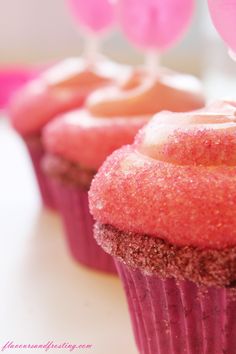 three pink cupcakes with frosting and balloons in the background