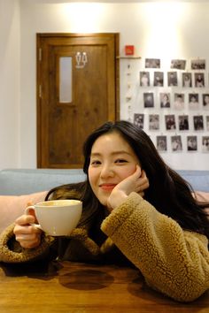 a woman sitting at a table with a coffee cup in her hand and looking into the camera