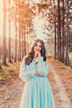 a woman in a blue dress is standing on a dirt road with her arms crossed