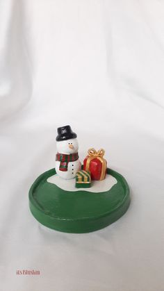 a small snowman sitting on top of a green plate next to a gift box